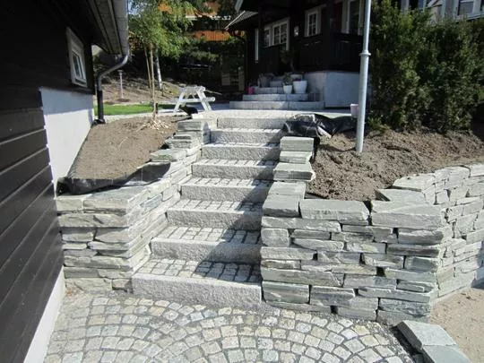 Brosteinsbelagt område og trapp, samt mur av flatstablede heller, ved garasje med mørkt malte panel.