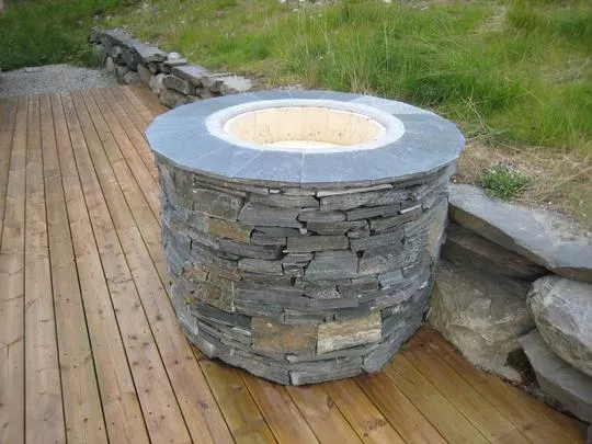 Rund, utepeis laget av stablet naturstein, plassert på en terrasse med tredekke og omkranset av gress.